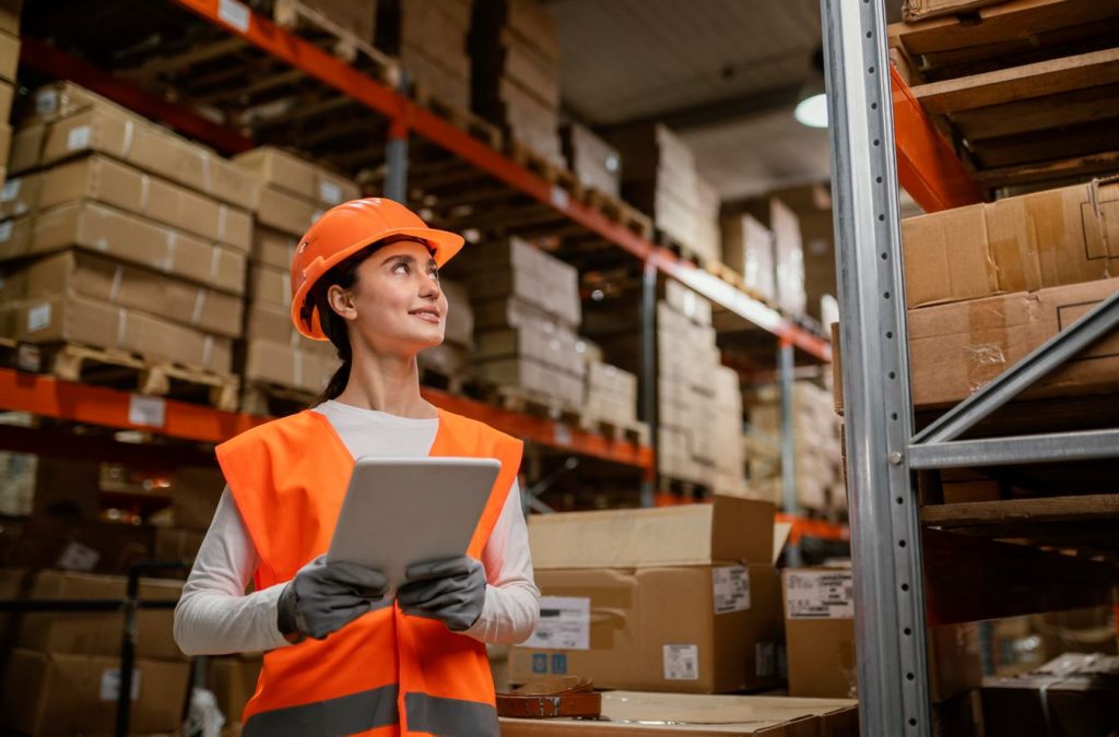Mulher em ambiente de estoque de produtos, vestindo equipamentos de EPI e segurando tablet.