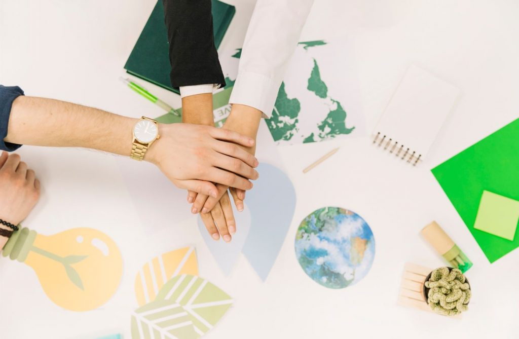 Vista de cima, temos as mãos de três pessoas em um sinal de acordo, união, posicionadas sobre uma mesa com recortes de símbolos que remetem à sustentabilidade