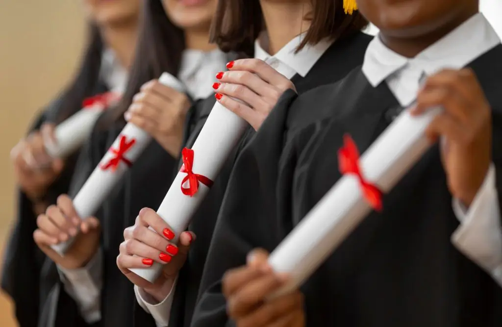 Foco nas mãos de quatro estudantes de beca segurando seus diplomas enrolados e presos com fitas