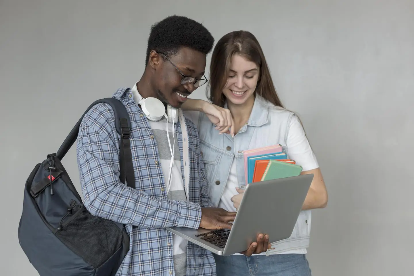 Dois estudantes jovens, sendo um rapaz e uma moça, olhando para um notebook. Ela segura alguns livros, e ele, uma mochila
