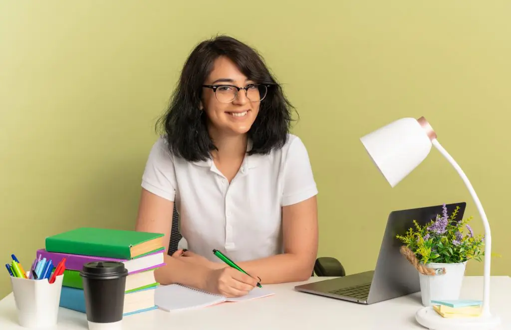 Mulher sorridente segura uma caneta e simula escrever um um caderno. Outros objetos estão sobre a mesa, como notebook, livros, luminária, copo e canetas