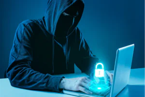 Homem encapuzado representando um hacker utiliza um notebook, no qual aparece um símbolo virtual de cadeado representando a segurança da informação