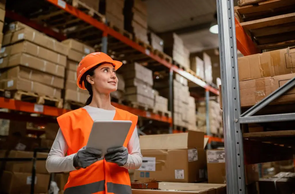 Mulher em ambiente de estoque de produtos, vestindo equipamentos de EPI e segurando tablet.